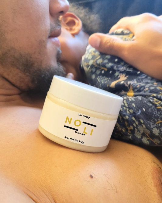 A jar of 'NOLI' men's deep hydration butter placed on a man's chest with a blurred image of a baby in the background.