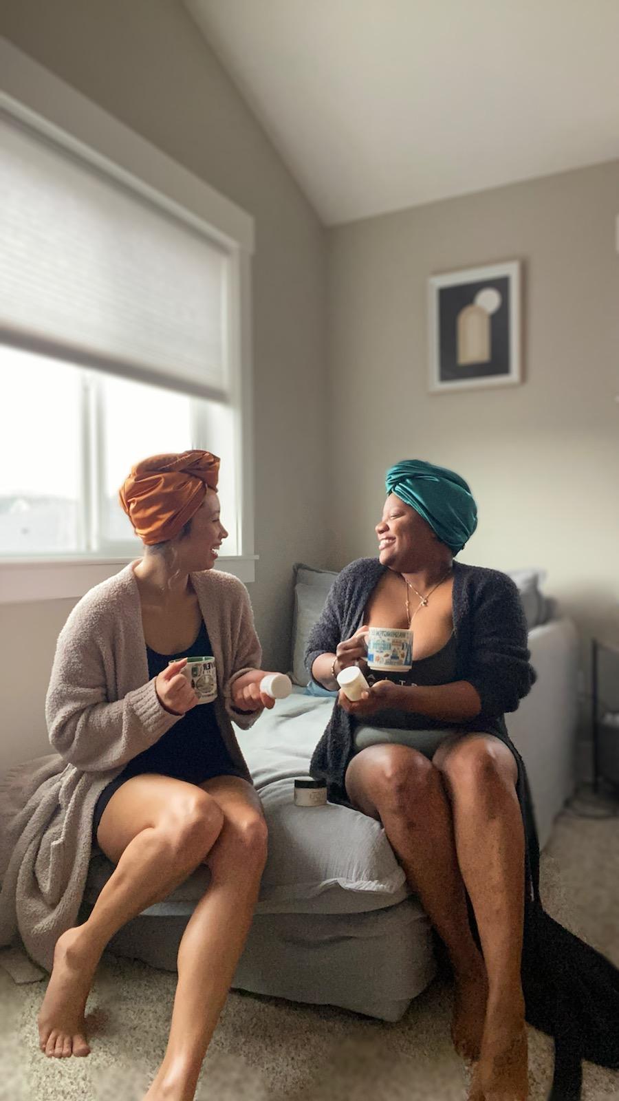 Two individuals seated on a sofa, wearing head wraps and engaging in a conversation while holding Noli Skin Care Hydration Butters