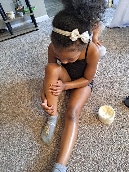 A child sitting and applying Kids Comfort Hydration Butter on her legs, with the butter container visible in the background.