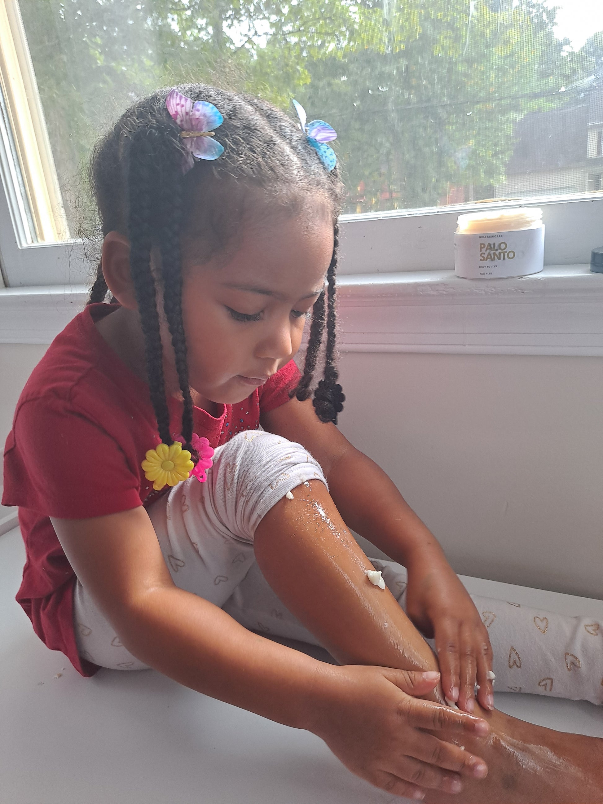 A child sitting and applying Kids Comfort Hydration Butter on her legs, with the butter container visible in the background.